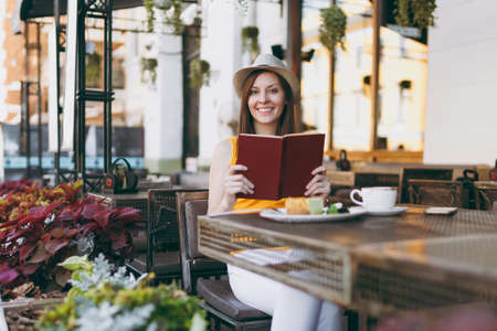 Woman in outdoors street coffee shop cafe sitting at table in hat, reading book with cup of cappuccino, cake, relaxing in restaurant during free time. Mobile Office in summer. Lifestyle rest conceptの写真素材