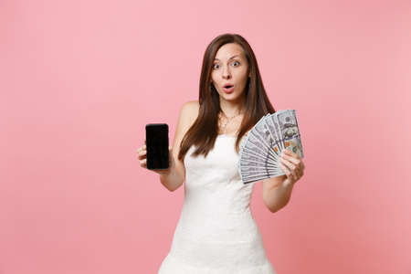 Shocked bride woman in wedding dress holding bundle lots of dollars cash money, mobile phone with...