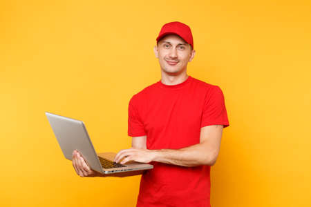 Delivery man isolated on yellow orange background. Professional male employee courier in red cap, t-shirt holding working typing on laptop pc computer. Service concept. Copy space for advertisementの写真素材