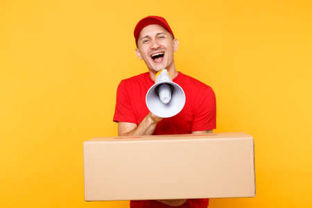 Delivery service man isolated on yellow background. Male employee courier in red cap, t-shirt scream in megaphone, hold empty cardboard box. Guy announces discounts sale. Receiving package conceptの写真素材