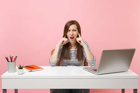 Young irritated grumpy woman does not want listen covering ears with finger sit work at white desk with contemporary pc laptop isolated on pink background. Achievement business career. Copy spaceの写真素材