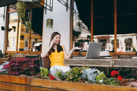 Happy girl in outdoors street cafe sitting at table with laptop pc computer, talking on mobile phone, conducting pleasant conversation, in restaurant during free time. Mobile office, freelance conceptの写真素材