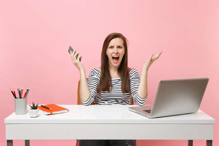 Young irritated woman spreading hands screaming holding mobile phone while sitting working at office with pc laptop isolated on pastel pink background. Achievement business career concept. Copy spaceの写真素材