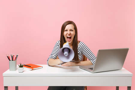 Irritated woman screaming in megaphone while sit and working on project at office with pc laptop isolated on pastel pink background. Achievement business career concept. Copy space for advertisementの写真素材
