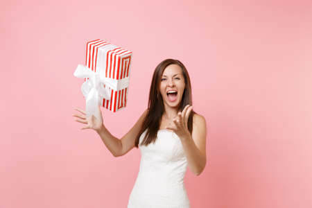 Funny joyful bride woman in elegant white wedding dress screaming throwing up red box with gift present isolated on pink pastel background. Wedding celebration concept. Copy space for advertisementの写真素材