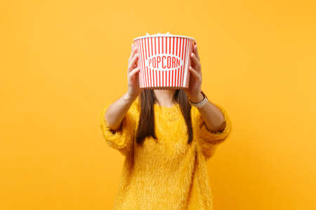 Young girl in fur sweater watching movie film and covering face with bucket of popcorn isolated on bright yellow background. People sincere emotions in cinema, lifestyle concept. Advertising areaの写真素材