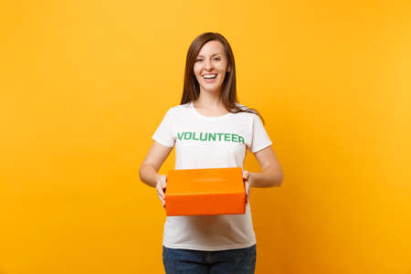 Portrait of smiling woman in white t-shirt with written inscription green title volunteer hold cardboard box isolated on yellow background. Voluntary free assistance help, charity grace concept.の写真素材