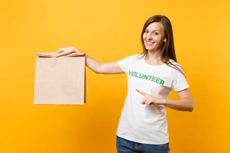 Portrait of woman in white t-shirt written inscription green title volunteer hold blank craft paper bag for takeaway isolated on yellow background. Voluntary free assistance charity grace conceptの写真素材