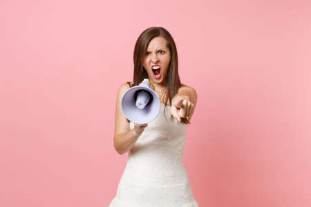 Aggressive angry bride woman in wedding dress screaming in megaphone, pointing index finger on camera isolated on pink pastel background. Organization of wedding concept. Copy space for advertisementの写真素材