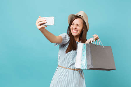 Portrait smiling woman in summer dress straw hat holding packages bags with purchases after shopping doing selfie shot on mobile phone isolated on blue pastel background. Copy space for advertisementの写真素材