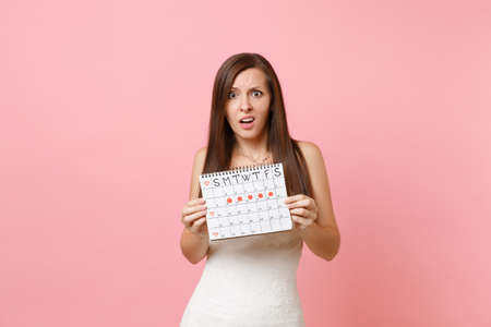 Shocked concerned bride woman in white wedding dress holding female periods calendar for checking menstruation days isolated on pink background. Medical, healthcare, gynecological concept. Copy spaceの写真素材