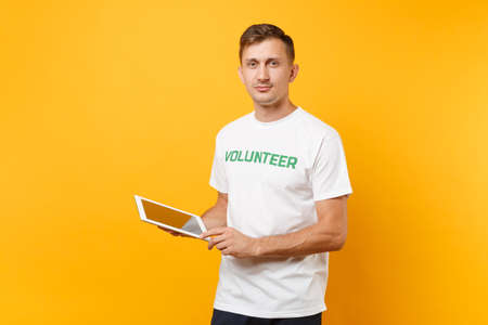 Portrait man in white t-shirt with written inscription green title volunteer using typing tablet pc computer isolated on yellow background. Voluntary free assistance help, charity grace work conceptの写真素材