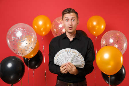 Shocked amazed young man in classic shirt holding bundle lots of dollars, cash money on red...
