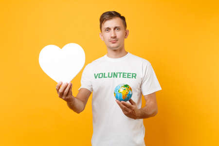 Portrait of man in white t-shirt with written inscription green title volunteer hold blank heart, Earth world globe isolated on yellow background. Voluntary free assistance help charity grace conceptの写真素材