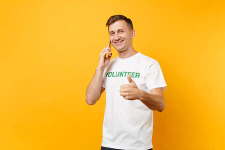 Young man portrait in white t-shirt with written inscription green title volunteer talking on mobile phone isolated on yellow background. Voluntary free assistance help, charity grace work conceptの写真素材