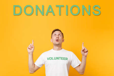 Man in white t-shirt with written inscription green volunteer looking up, pointing fingers on title donations isolated on yellow background. Voluntary free assistance help, charity grace work conceptの写真素材