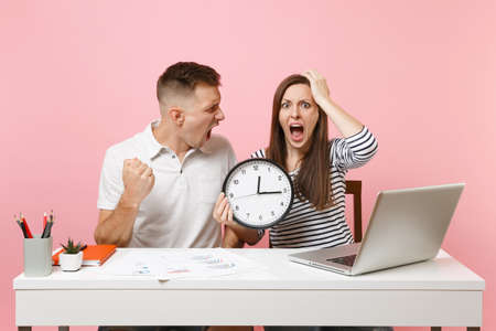 Two young fun shocked business woman man colleagues sit work at white desk with clock, laptop...