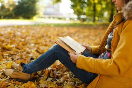 The woman in yellow coat and jeans sitting under the maple tree with a red book in fall city park on...