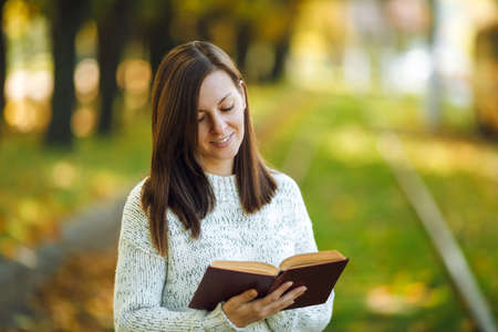 The beautiful happy smiling brown-haired woman in white sweater standing with a red book on the tram...