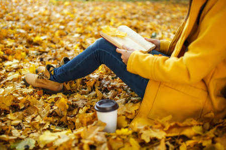 The woman in yellow coat, jeans and boots sitting under the maple tree with a red book and cup of coffee or tea in fall city park on a warm day. Autumn golden leaves. Reading concept. Close up.の写真素材