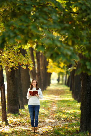 The beautiful happy smiling brown-haired woman in white sweater standing with a red book in fall city park on a warm day. Autumn golden leaves. Reading concept.の写真素材