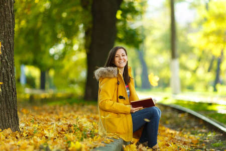 The beautiful happy brown-haired woman in a yellow coat and jeans sits alone in the park near the...