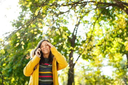 The beautiful happy cheerful brown-haired woman in a yellow coat and striped longsleeve talking on mobile phone in fall city park on a warm day. Autumn golden leaves.の写真素材
