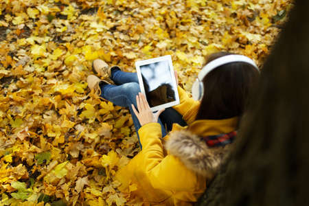 The brown-haired woman in yellow coat and jeans sitting and listening to music under a tree with a tablet in her hands and headphones in fall city park on a warm day. Autumn golden leaves. Top viewの写真素材