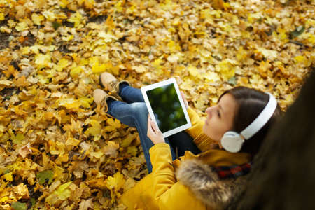 The brown-haired woman in yellow coat and jeans sitting and listening to music under a tree with a tablet in her hands and headphones in fall city park on a warm day. Autumn golden leaves. Top viewの写真素材