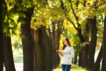 The beautiful happy smiling brown-haired woman in white sweater standing with a red book in fall...