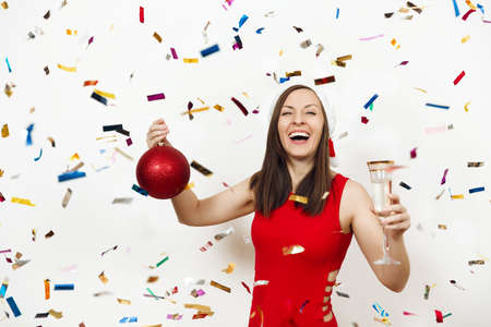 Beautiful smiling caucasian young happy woman in red dress and Christmas hat standing with big tree toy and glass of champagne on white background confetti. Santa girl isolated. New Year 2018 holiday.の写真素材