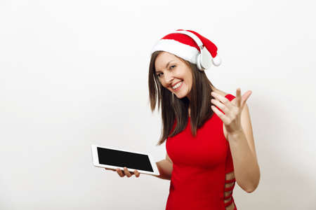 Pretty young happy woman in red dress and Christmas hat listening music on headphones, holding tablet on white background. Santa girl gadget isolated. New Year holiday 2018. Copy space advertisementの写真素材