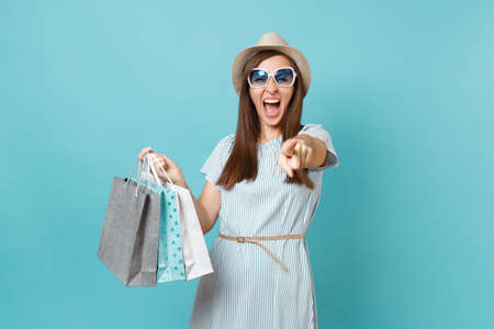 Portrait fashionable attractive happy woman in summer dress, straw hat, sunglasses holding packages bags with purchases after shopping isolated on blue pastel background. Copy space for advertisementの写真素材