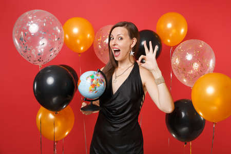 Excited young woman in black dress celebrating, holding world globe, showing OK sign on bright red background air balloons. St. Valentine's Day, Happy New Year, birthday mockup holiday party conceptの写真素材