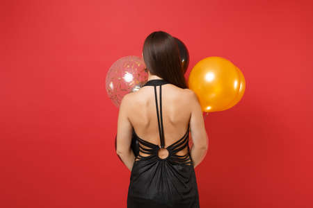 Back rear view of young woman in black dress holding air balloons, celebrating isolated on red...