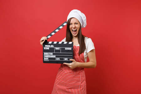 Housewife female chef cook or baker in striped apron, white t-shirt, toque chefs hat isolated on red wall background. Woman holding classic black film making clapperboard. Mock up copy space conceptの写真素材