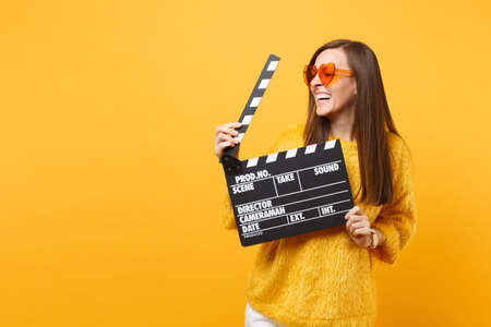 Laughing young woman in orange heart eyeglasses looking aside and holding classic black film making clapperboard isolated on yellow background. People sincere emotions, lifestyle. Advertising areaの写真素材
