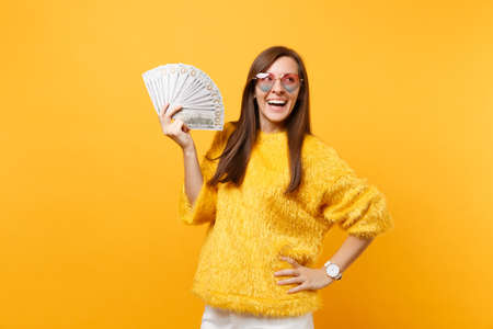 Laughing young woman in heart eyeglasses looking up and holding bundle lots of dollars, cash money...
