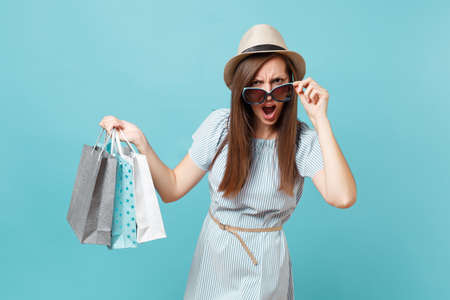 Portrait fashionable attractive happy woman in summer dress, straw hat, sunglasses holding packages bags with purchases after shopping isolated on blue pastel background. Copy space for advertisementの写真素材
