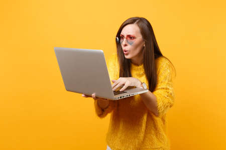 Portrait of shocked young woman in fur sweater and heart eyeglasses working on laptop pc computer isolated on bright yellow background. People sincere emotions, lifestyle concept. Advertising areaの写真素材