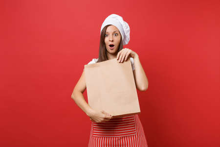 Housewife female chef cook baker in striped apron t-shirt toque chefs hat isolated on red wall background.Woman hold in hand brown clear blank craft paper bag takeaway Mock up copy space conceptの写真素材