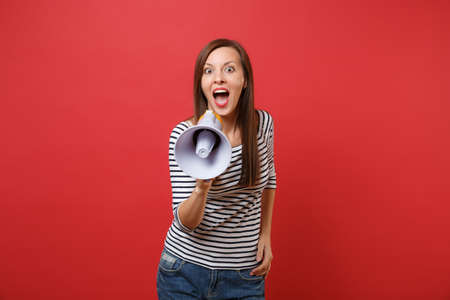 Excited young woman in striped clothes screaming on megaphone keeping mouth wide open looking...