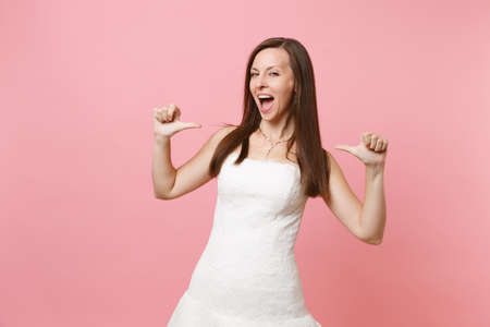 Portrait of narcissistic bride woman with opened mouth in elegant lace white wedding dress pointing thumbs on herself isolated on pastel pink background. Wedding concept. Copy space for advertisementの写真素材