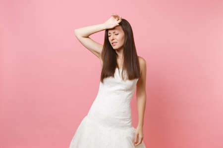 Portrait of exhausted bride woman in white wedding dress keeping hand on forehead tired of preparing for wedding isolated on pink background. Wedding celebration concept. Copy space for advertisementの写真素材