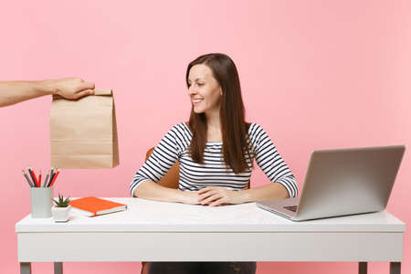 Woman looking on brown clear empty blank craft paper bag, work at desk with pc laptop isolated on pink background. Food products delivery courier service from shop or restaurant to office. Copy spaceの写真素材