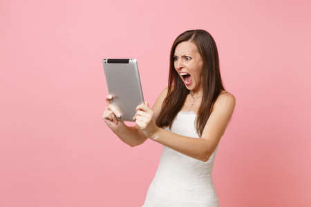 Irritated angry bride woman in wedding dress screaming swearing working on tablet computer making video call isolated on pink background. Wedding to do list. Organization of celebration. Copy spaceの写真素材