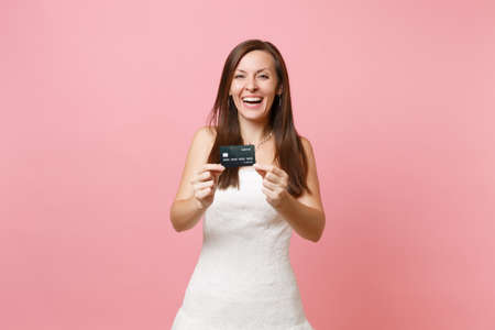 Portrait of laughing bride woman in white wedding dress showing credit card on camera isolated on pink pastel background. Organization of wedding celebration concept. Copy space for advertisementの写真素材