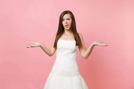 Portrait of concerned upset bride woman in elegant lace white wedding dress standing and spreading hands isolated on pastel pink background. Wedding celebration concept. Copy space for advertisementの写真素材
