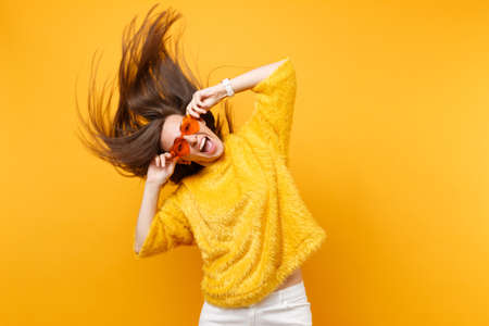 Excited young girl in fur sweater holding heart orange glasses fooling around in studio jumping with fluttering hair isolated on yellow background. People sincere emotions lifestyle. Advertising areaの写真素材