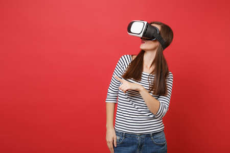 Young woman in casual striped clothes virtual reality glasses pointing index finger looking aside...